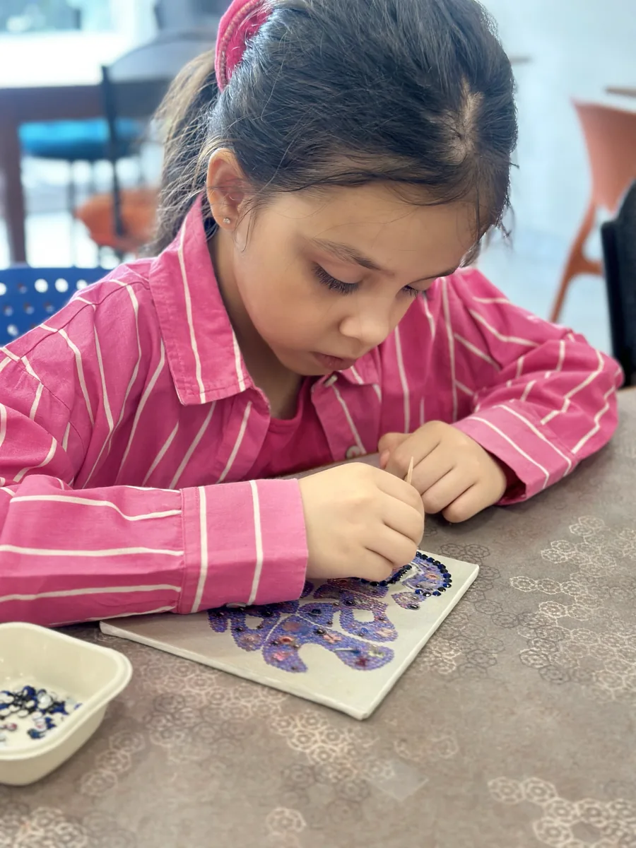 A young girl placing tiny beads onto a small painted canvas with a toothpick, concentrating closely